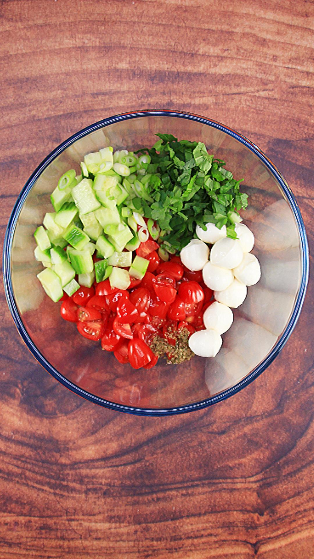 Ingredients in a bowl before they are stirred: cucumber, parsley, basil, tomatoes, and fresh mozzarella, with oregano, salt and pepper.