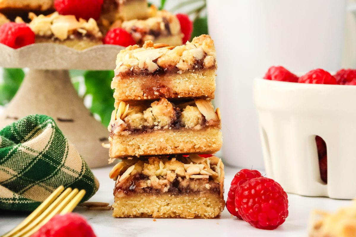 A side view of a stack of three raspberry bars made with a shortbread crust, raspberry jelly, and a crumble top. Hostess At Heart