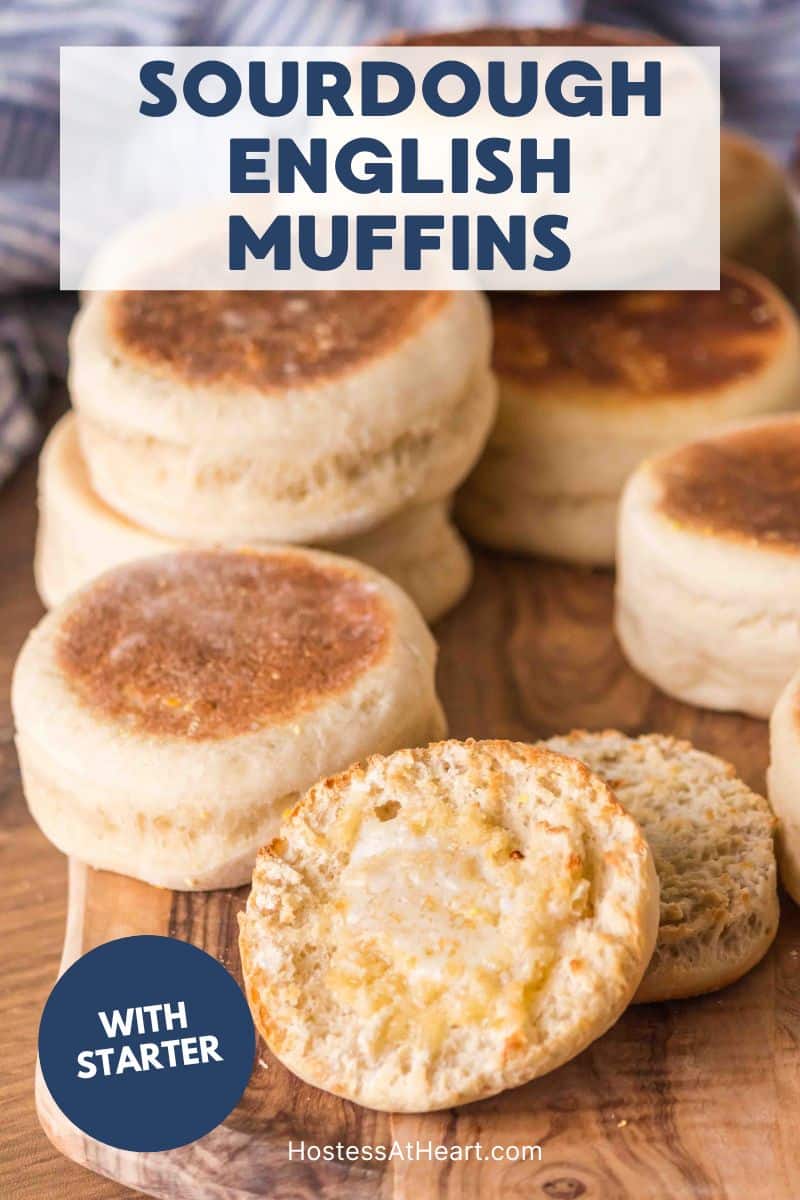 A batch of homemade Sourdough English Muffins sitting on a cutting board. The from english muffin is split in half and buttered. Hostess At Heart
