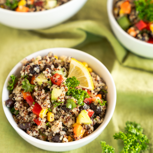 Top down view of three bowls filled with a mixture of tri-color quinoa and chopped fresh veggies including cucumber, carrots, corn, and red peppers, garnished with lemon and parsley.