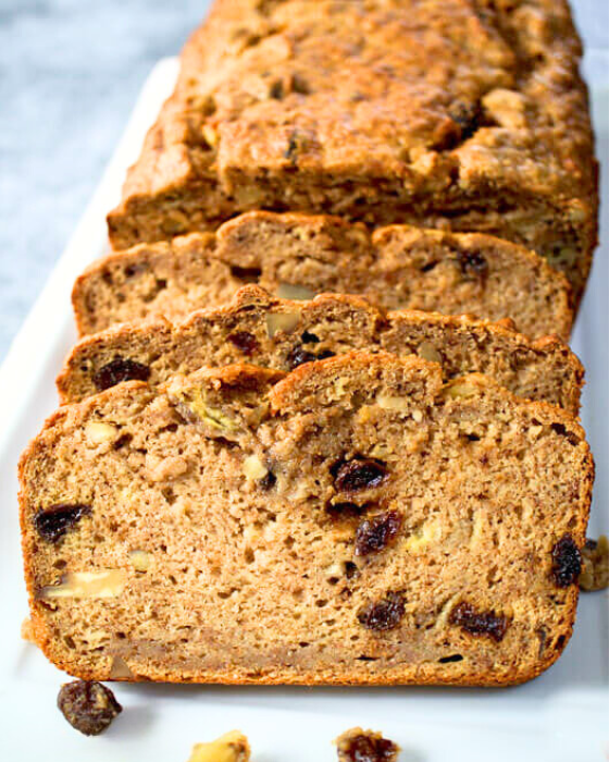 A loaf of quick apple banana bread dotted with raisins and chopped pecans on a plate with the front 3 slices fanned out in the front. Hostess At Heart