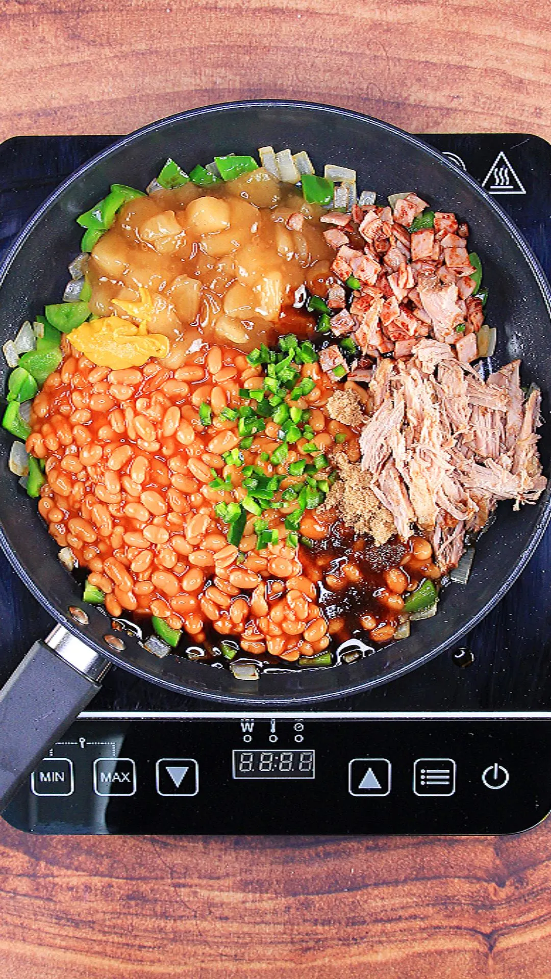 A skillet filled with ingredients: baked beans, pulled pork, apple pie filling, onion, green pepper, and bacon.