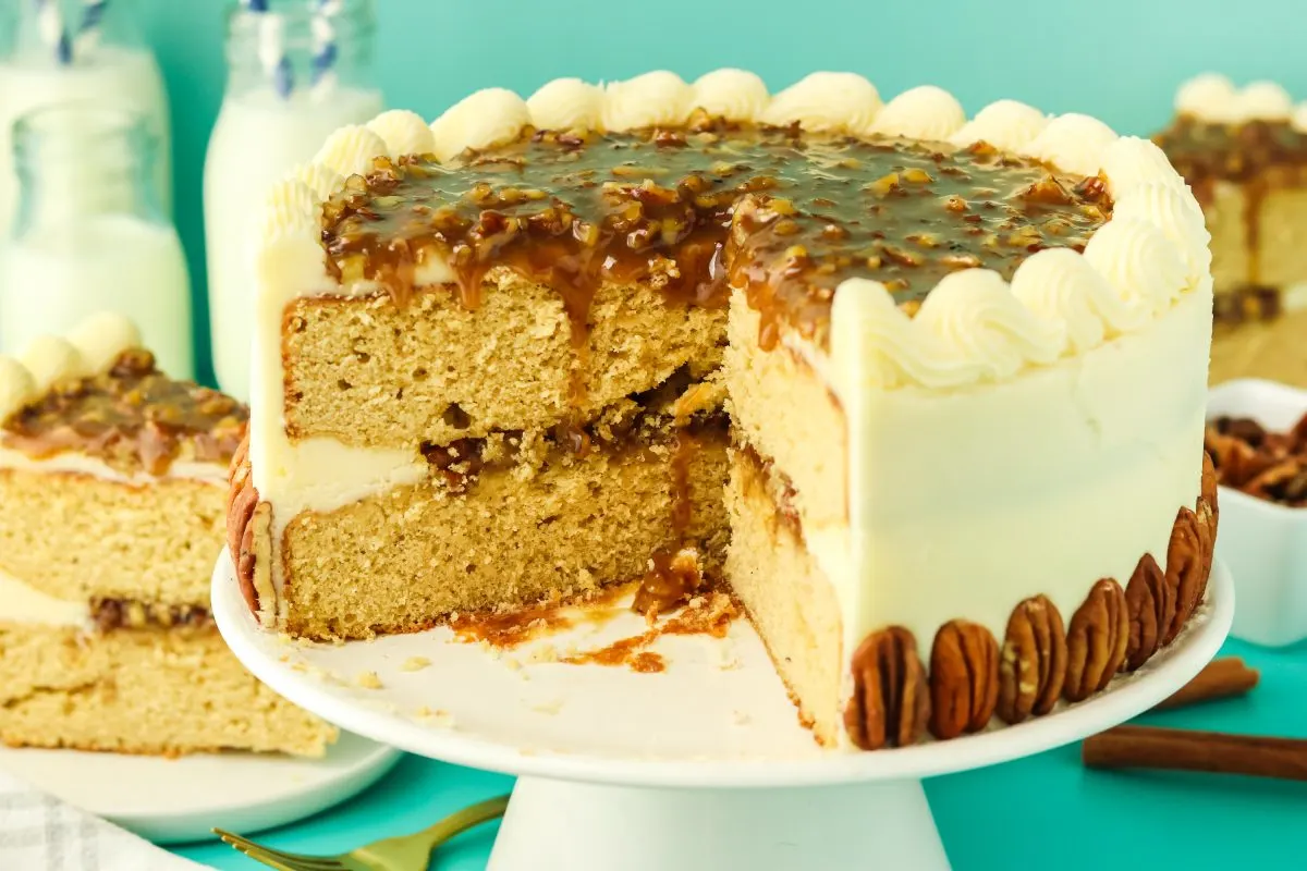 A round pecan layer cake with a large slice removed showing a pecan filling and buttercream frosting between the cake layers. The sides are frosted with buttercream and garnished with whole pecans. The top pecan praline is dripping down the cut edge of the cake. Hostess At Heart