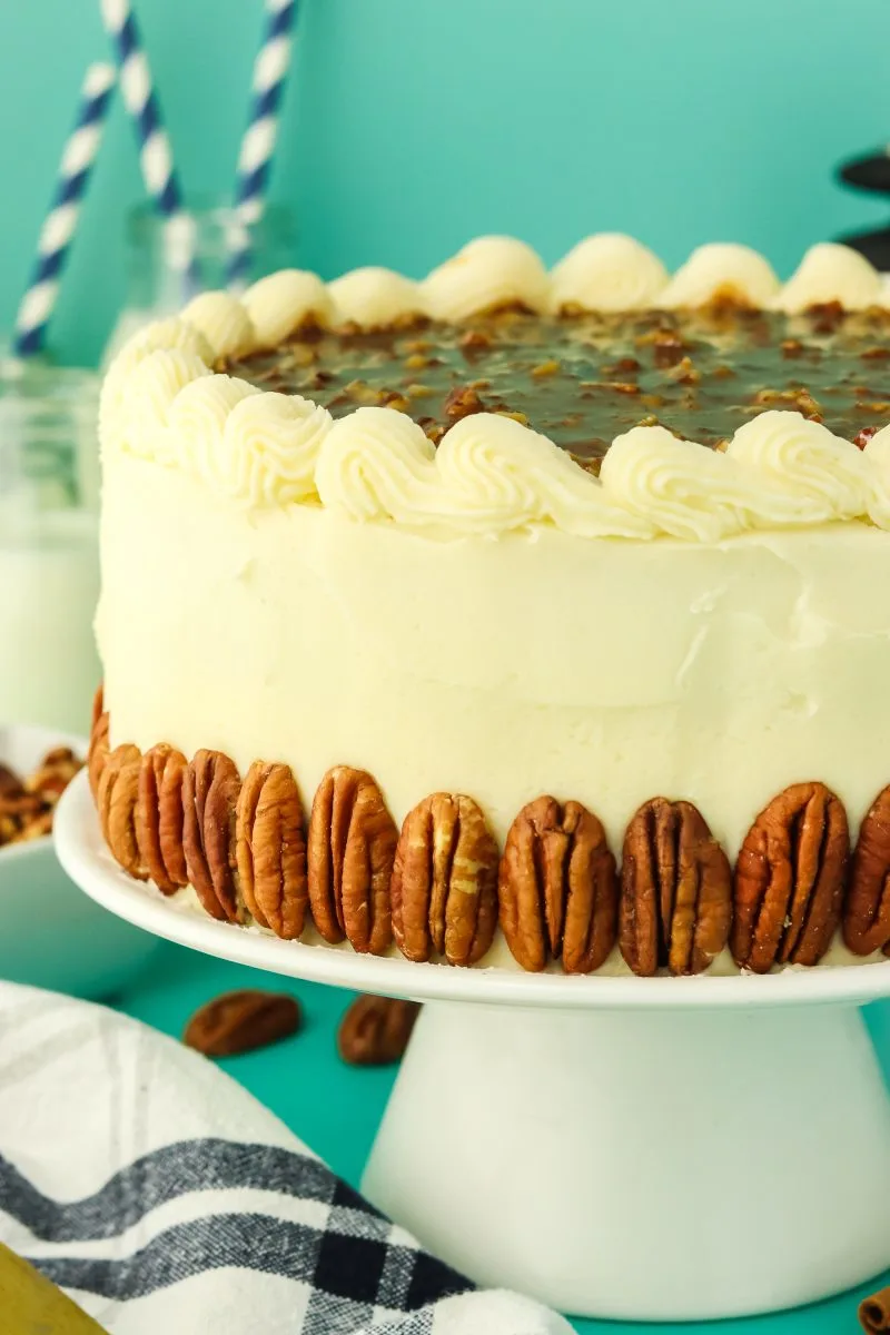 3/4 view of a round cake on a stand that's been frosted in butter cream, garnished with whole pecans and the top is layered with pecan filling. Hostess at Heart