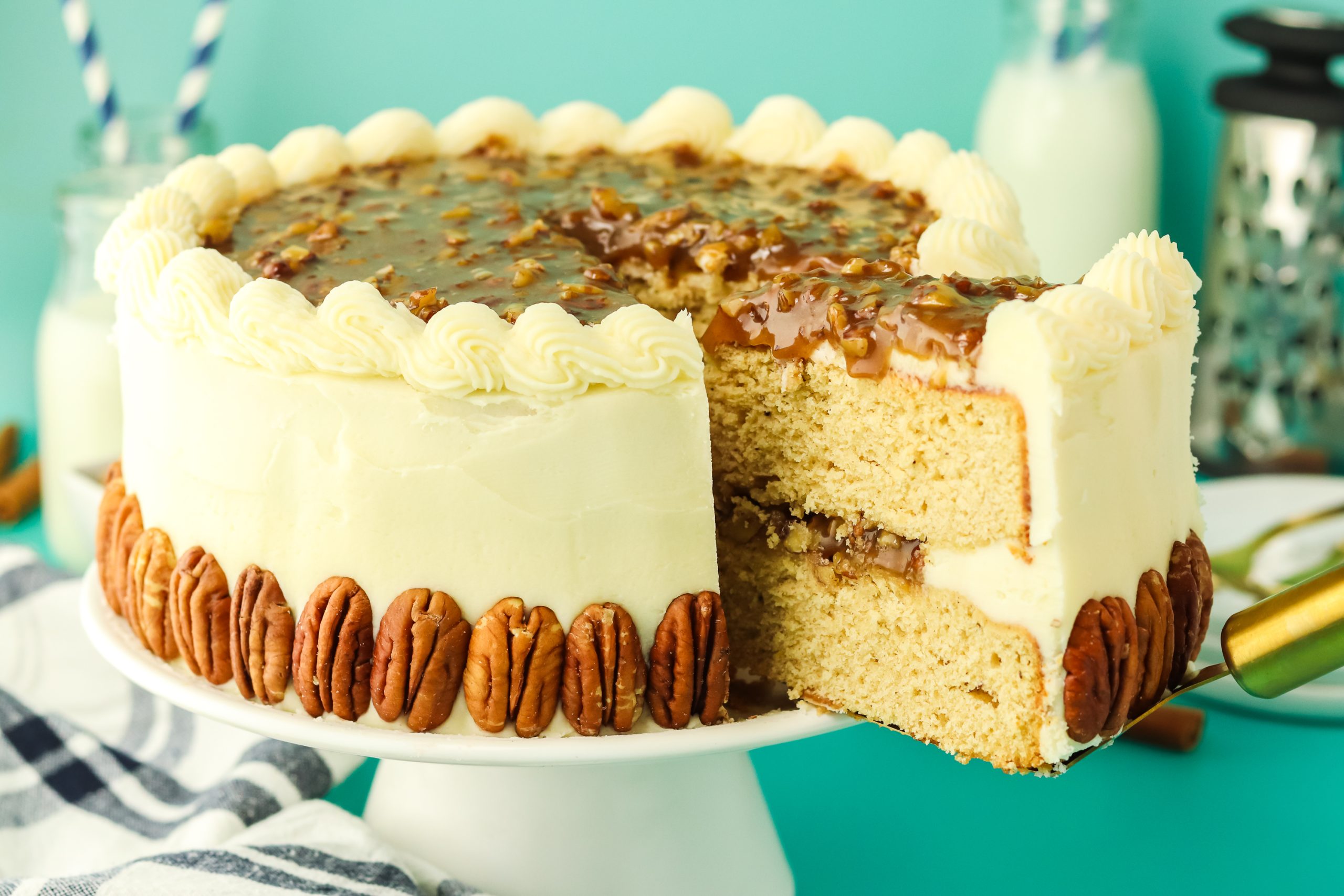 A round 2-layer cake with a slice being lifted out on a spatula showing a pecan filling between the two layers and frosted on the sides and garnished the bottom edge with whole pecans. Hostess At Heart