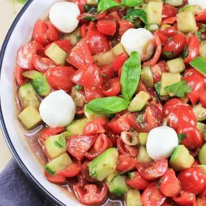 A close up top down view of a bowl of Caprese salad made of cherry tomatoes, pearl mozzarella balls, herbs, cucumber and dressed in a balsamic glaze.