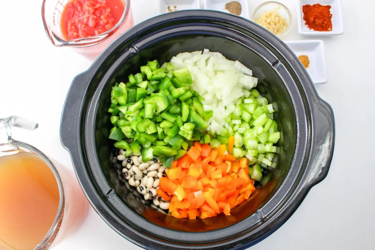 Top down view of a slow cooker filled with rice, chopped bell peppers, celery, and soaked black eyed peas. Hostess At Heart