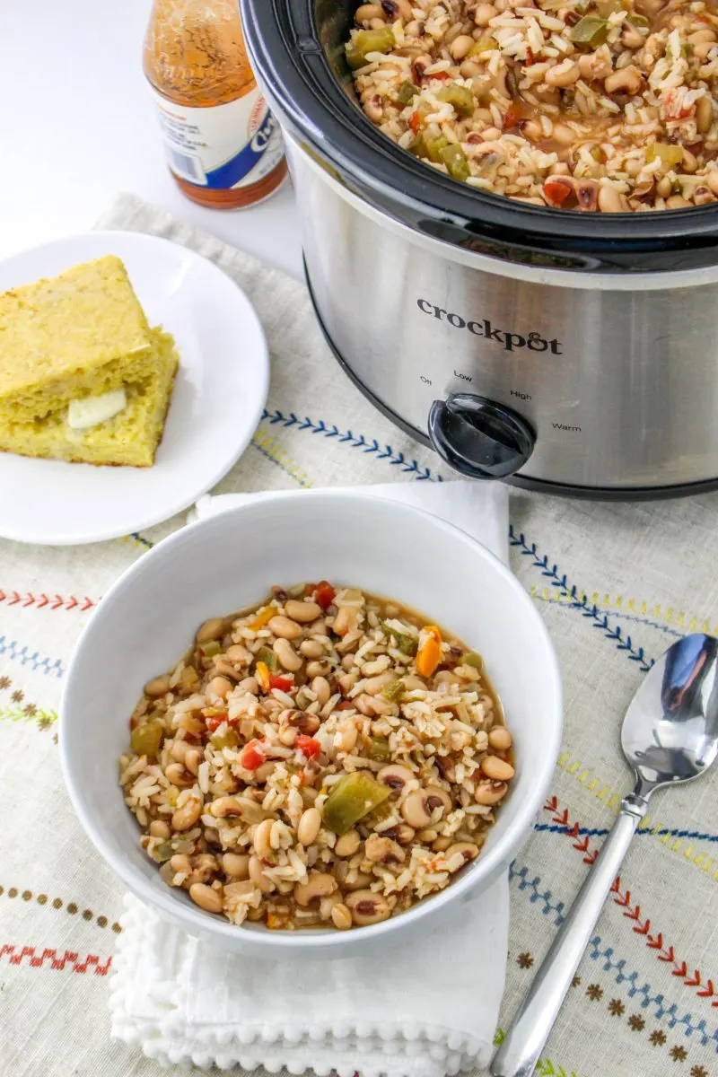 Top down view of a slow cooker and a bowl filled with cooked black-eyed peas and rice next to a plate of corned bread. Hostess At Heart