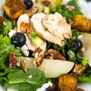 Angled view of Top down view of a salad of greens topped with sauteed pears, pecans, blueberries, slices of chicken, goat cheese crumbles and croutons - Hostess At Heart