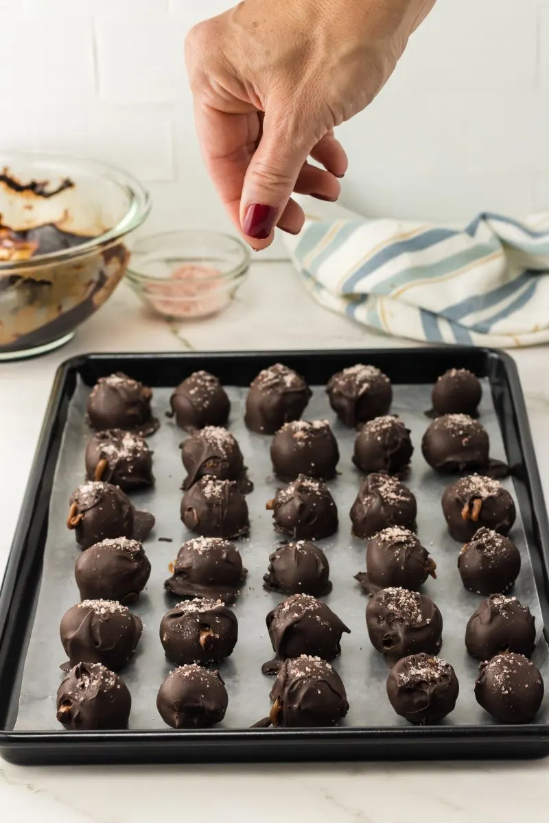 A hand sprinkling sea salt onto shaped caramel truffles on a sheet pan.