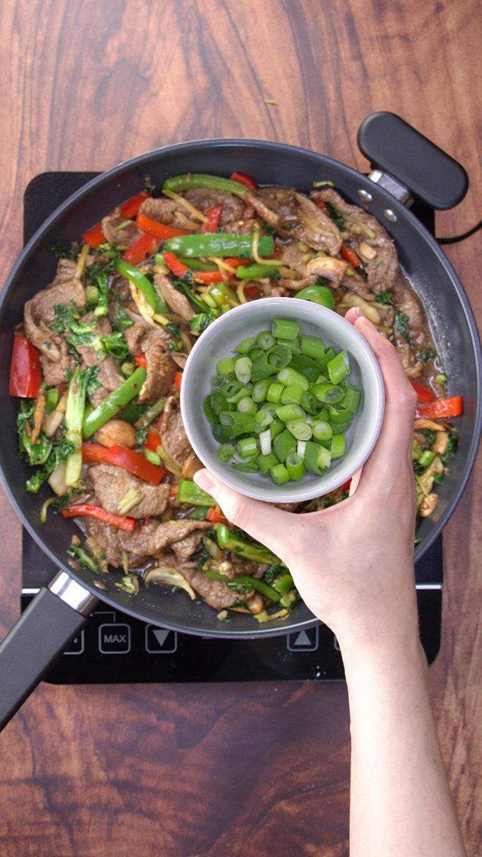 A hand holding a bowl of chopped green onions hovering over a skillet of beef stir fry.