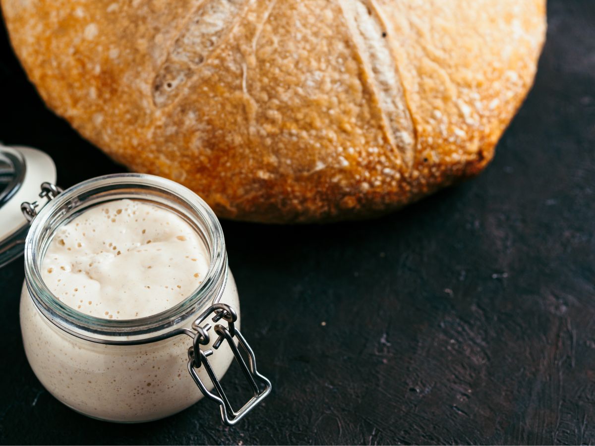 A jar of bubbly sourdough bread starter sitting next to a baked loaf of sourdough - Hostess At Heart