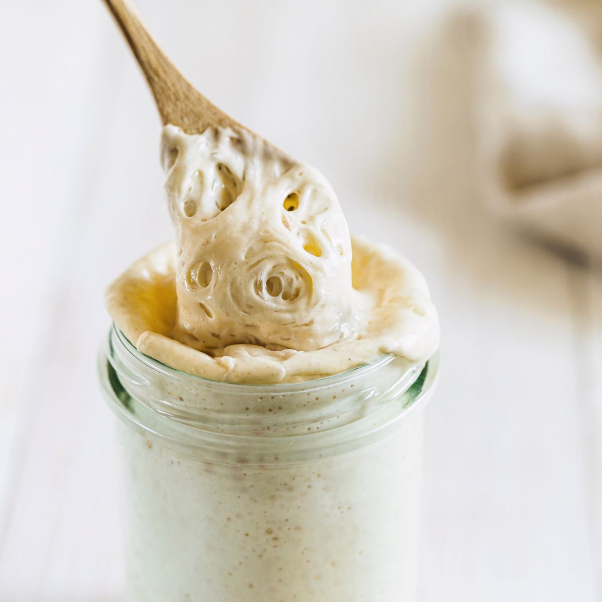 A wooden spoon pulling thick active sourdough starter out of a jar - Hostess At Heart