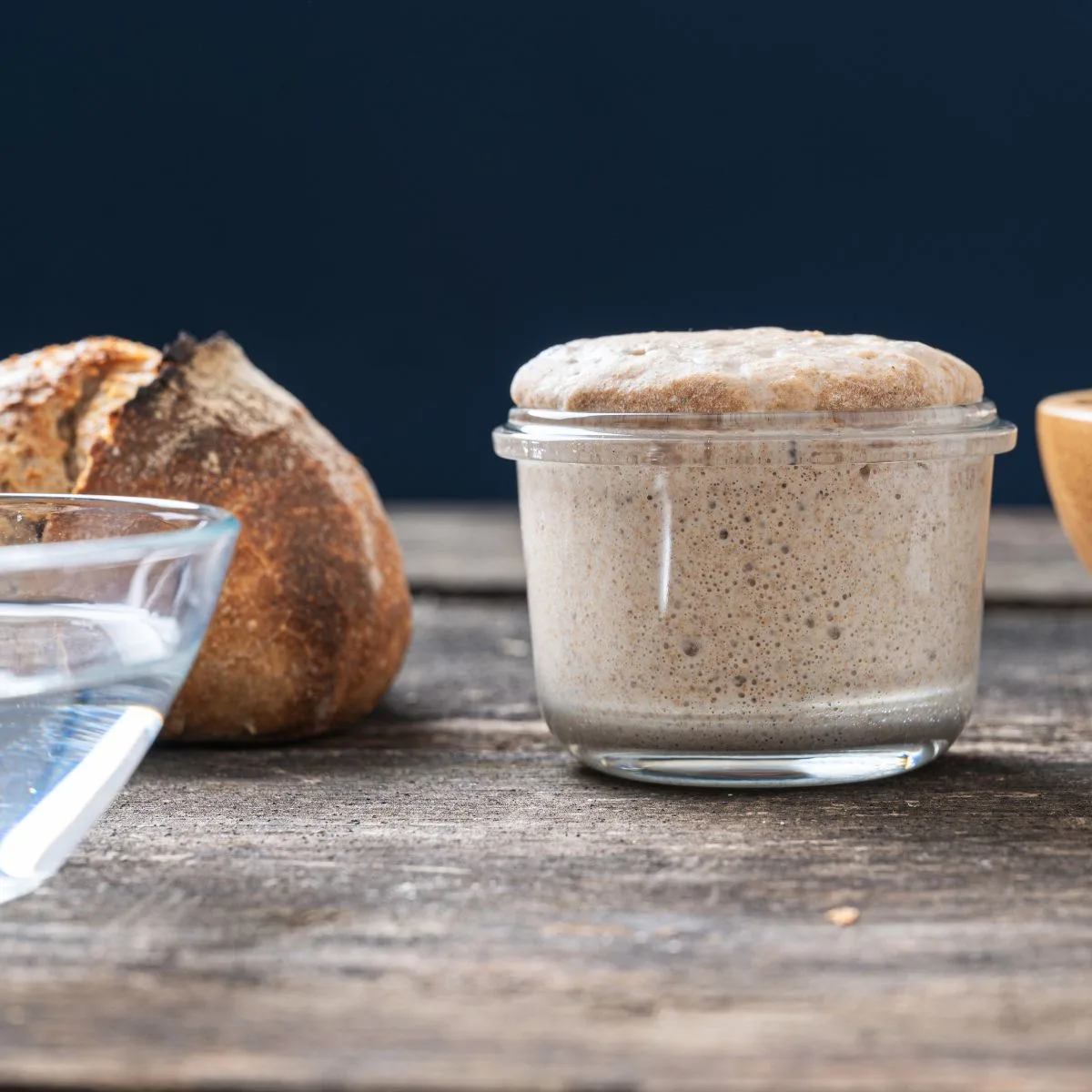 Sideview of a bubbly sourdough starter sitting next to a baked loaf of bread - Hostess At Heart