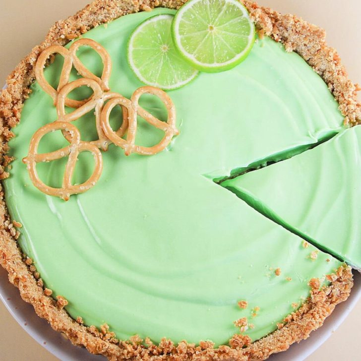 A top-down view of a green-colored cream pie with a pretzel crust topped with pretzels and lime wedges - Hostess At Heart