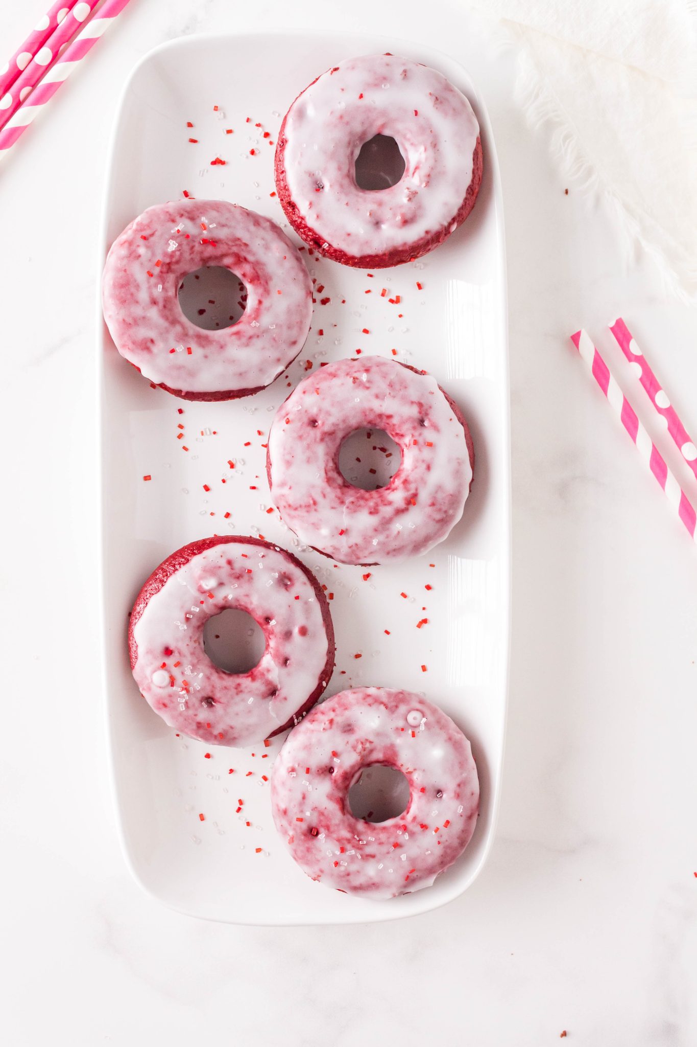 Top down view of a white plate filled with baked red velvet Valentine's Day donuts glazed and sprinkled. Hostess At Heart