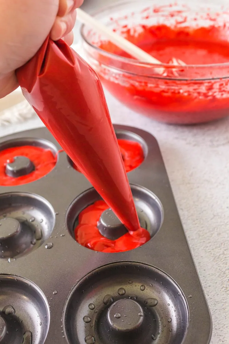 Piping donut batter into the pan with a mixing bowl full of red batter in the background.