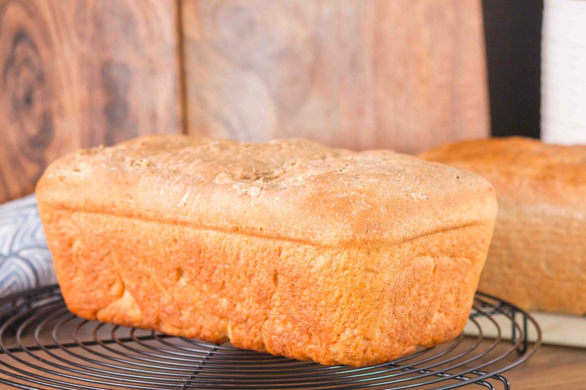 Full loaf of rye bread, side view, on a cooling rack. A second loaf is out of focus in the background.