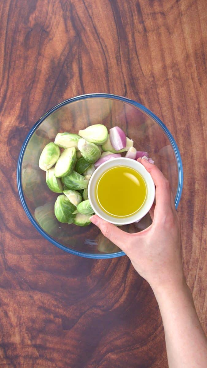 A bowl of olive oil hovering over a bowl of halved Brussel Sprouts.