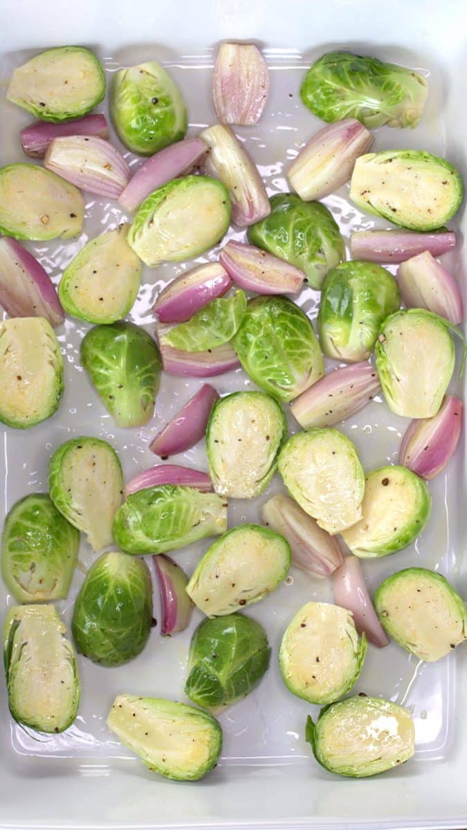 Raw halved Brussels sprouts and shallots covered in olive oil and salt and pepper in a baking dish.