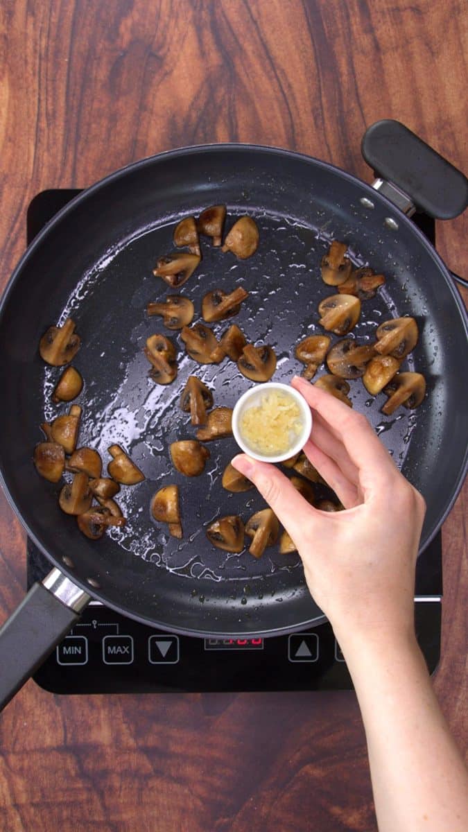 A bowl of minced garlic added to a skillet containing sauteed mushrooms.