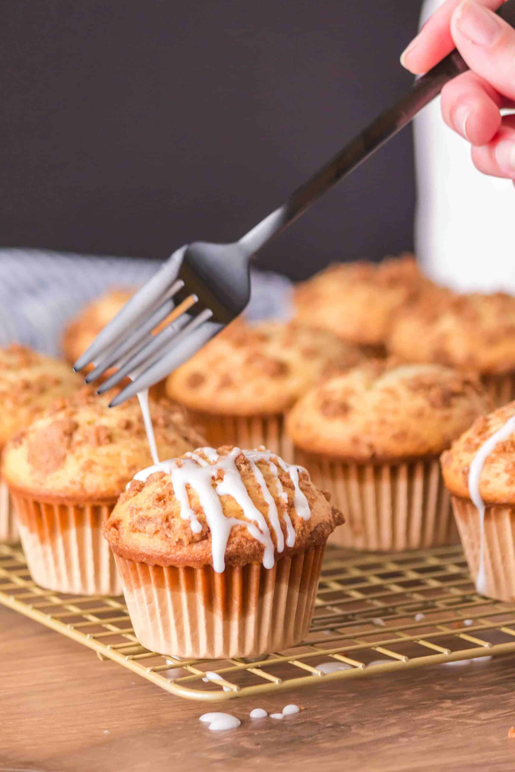 Angled view of a cinnamon streusel muffin sitting on a cooling rack being drizzled with a forkful of glaze. Hostess At Heart