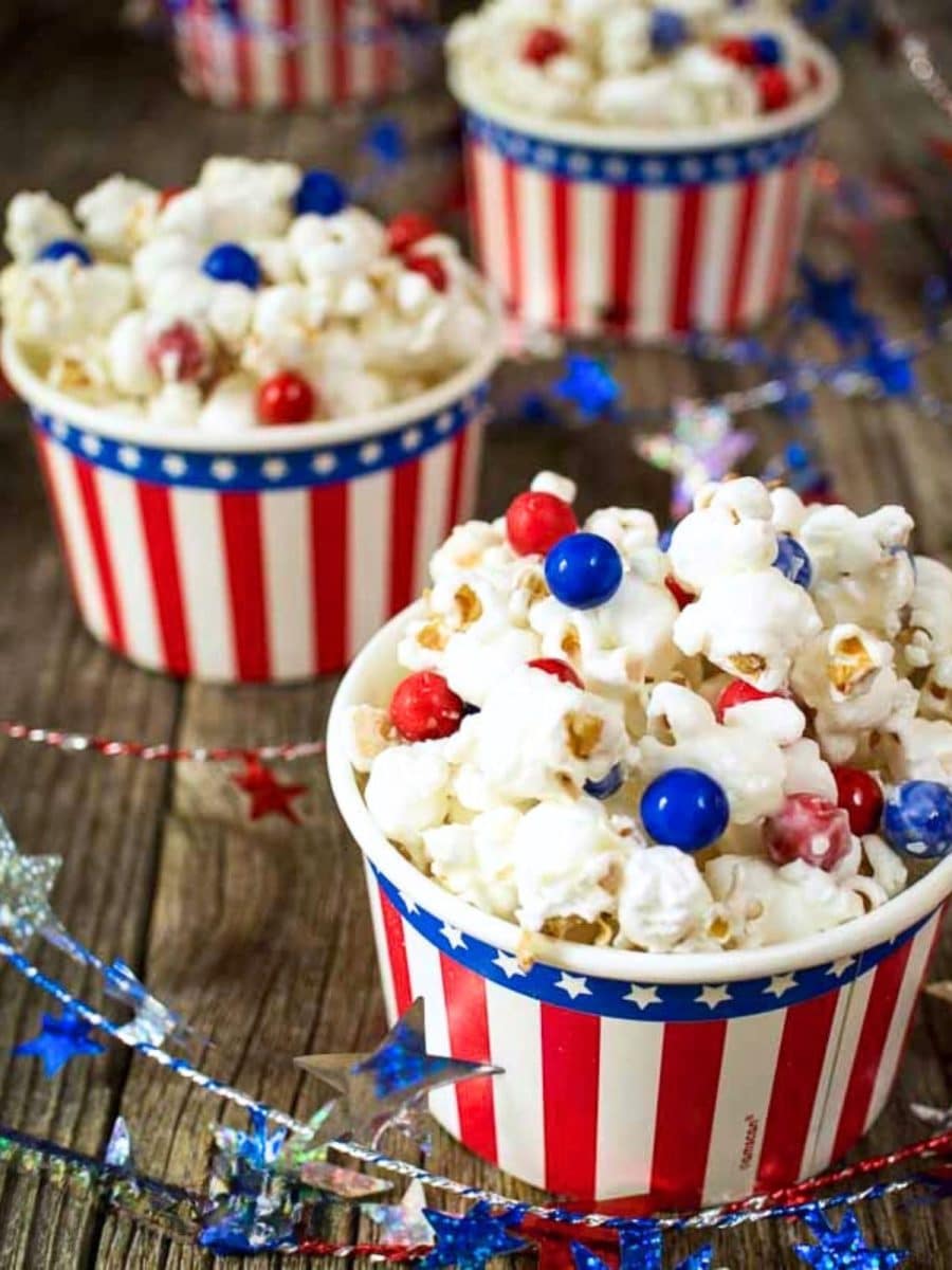 A sideview of a decorative cup holding pop corn with almond bark coating with red and blue candy pieces Hostess At Heart