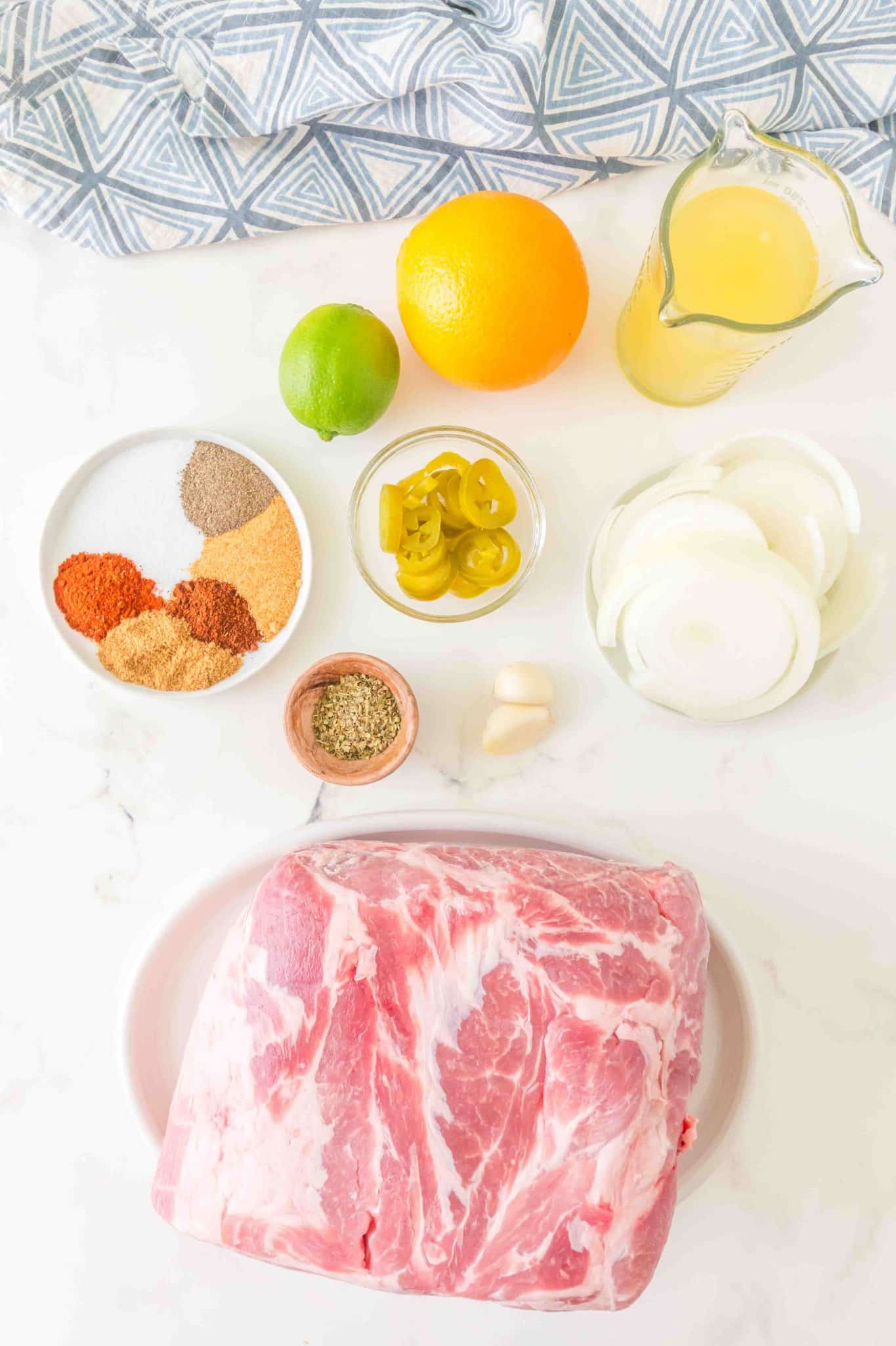 Top down view of ingredients: Pork shoulder, jalapeno peppers, onions, lime, orange, garlic, chicken broth, and ground spices. Hostess At Heart