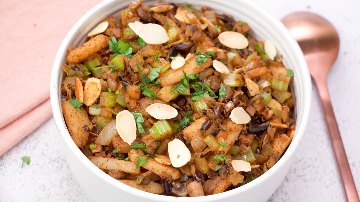 Angled view of a bowl of a mixture of 5 spice seasoned cooked rice topped with slices of almonds and chopped parsley. Hostess At Heart