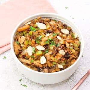 Top down view of a white bowl filled with cooked wild rice, celery, onion, mushrooms, chestnuts, and spices. It's Garnished with sliced almonds and parsley. Hostess At Heart