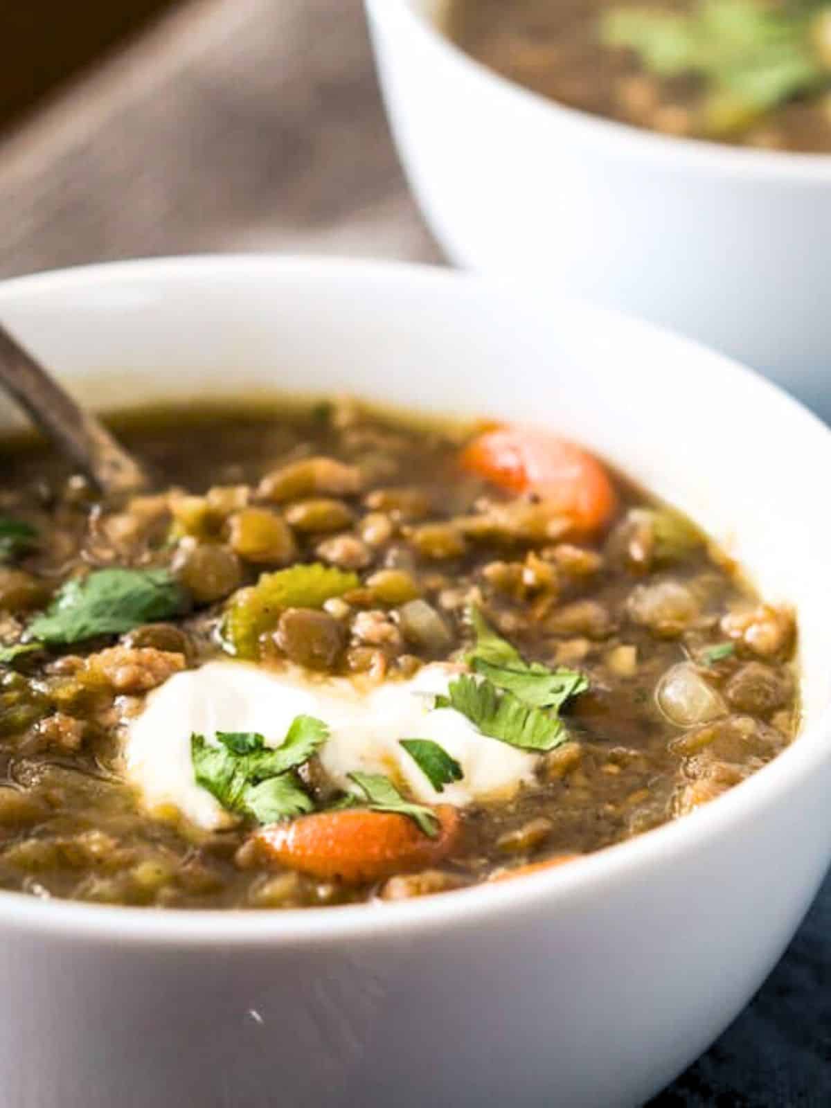 Closeup view of a bowl of ground sausage and lentil soup filled with vegetables. It's finished with a spoonful of yogurt and fresh cilantro. Hostess At Heart