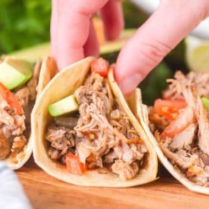 A tableview image of a hand grabbing a pork carnitas taco filled with avocado, onion, and tomato - Hostess At Heart