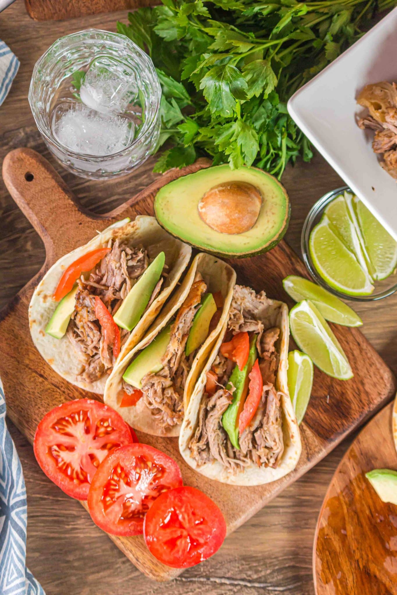 A top down view of three carnitas tacos on a cutting board surrounded by tomatoes, lime wedges, and an avocado. Hostess At Heart