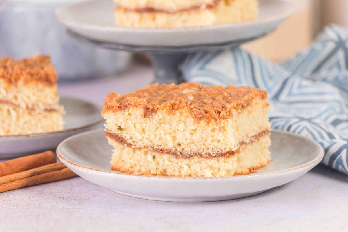Individual serving of Sourdough Discard Coffee Cake Recipe on a plate, with the remaining cake on a cake platter in the background, but blurry. Beautiful blue napkin also in the bakcground.