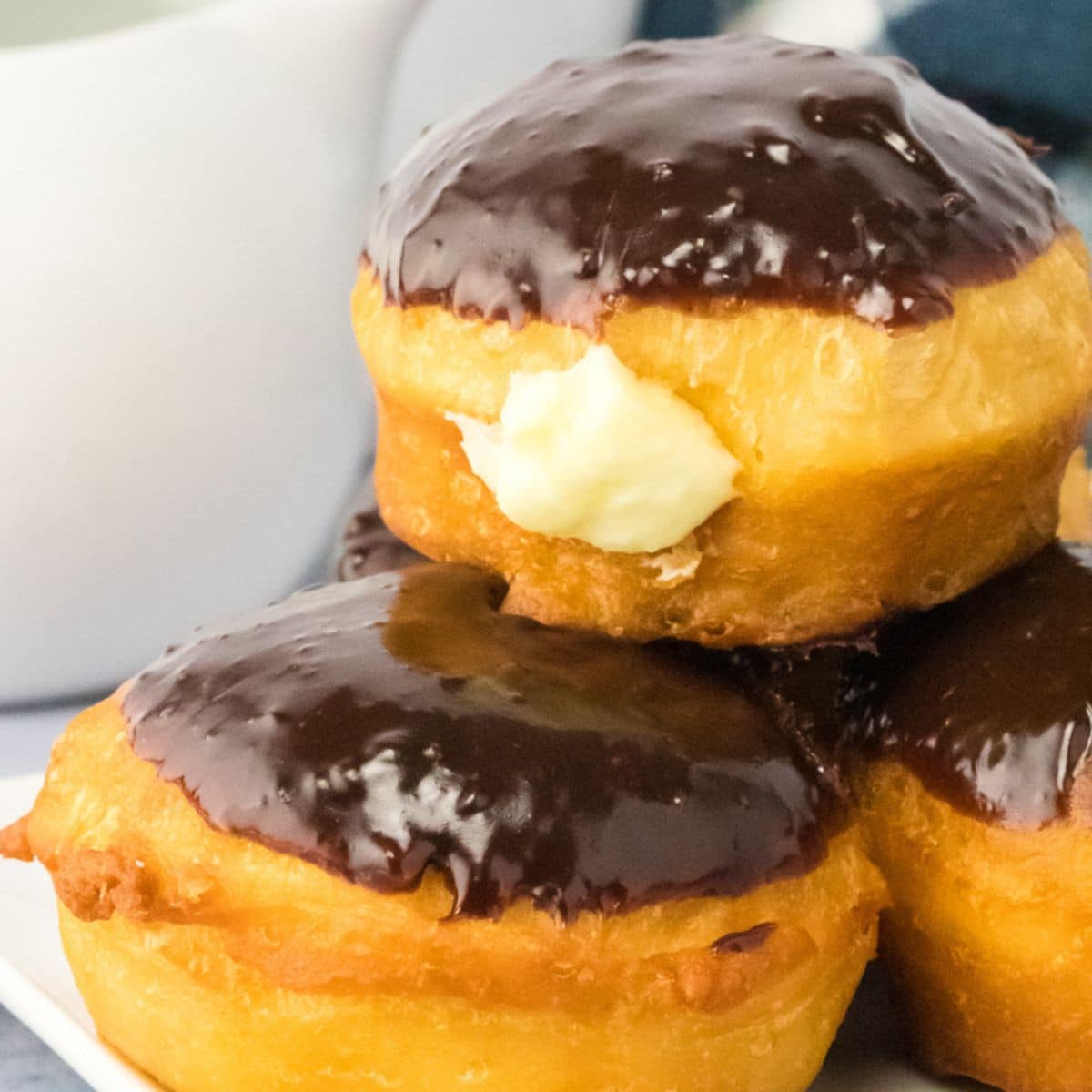 An image for Pinterest of stacked Boston Cream Donuts topped with chocolate ganache and with the cream filling oozing out of the side. Hostess At Heart