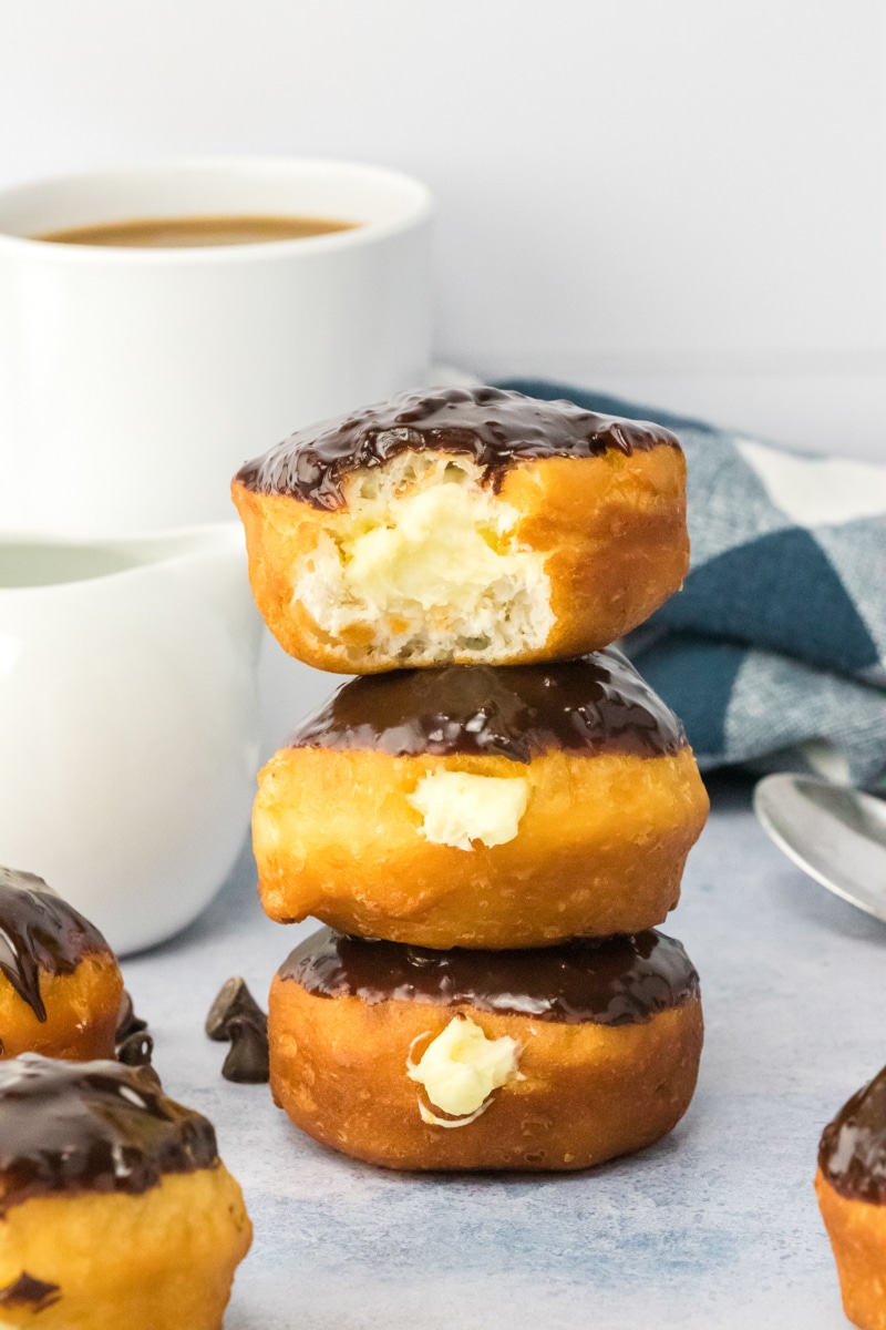 A stack of 3 Boston Cream Donuts with the top one having a bite taken out of it showing the cream filling. Hostess At Heart
