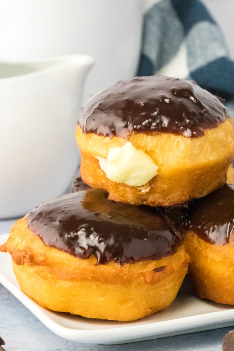 An image for Pinterest of stacked Boston Cream Donuts topped with chocolate ganache and with the cream filling oozing out of the side. Hostess At Heart