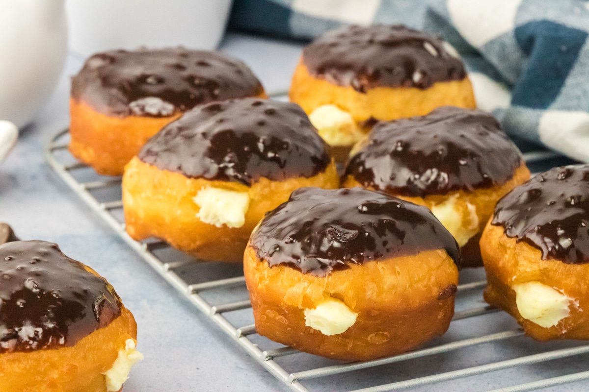 Sideview of Boston Cream Donuts sitting on a rack showing the cream coming out of the side and the tops have a shiny chocolate ganache. Hostess At Heart