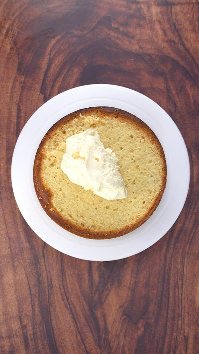 An image showing how to assemble a simple vanilla layer cake with the top sliced flat and a dollop of vanilla frosting waiting to be spread - Hostess At Heart
