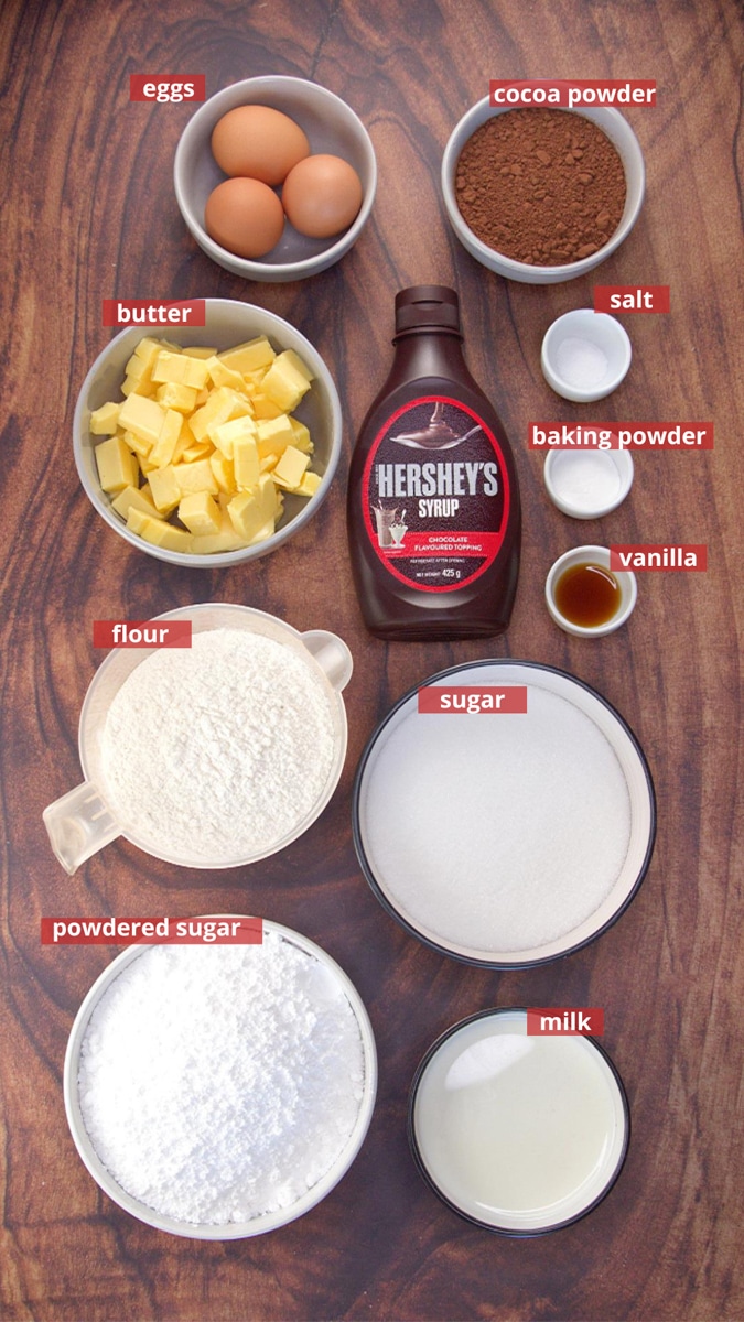 Ingredients used to make a glazed chocolate syrup cake including: flour, butter, sugar, Hershey Syrup, eggs, vanilla, baking powder, confectioners sugar, cocoa, and salt.