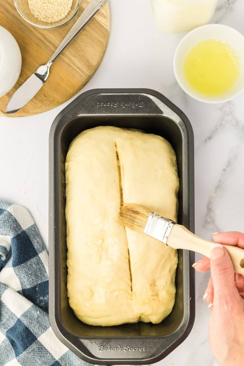 Egg wash being applied to a loaf of scored sesame seed dough in a loaf pan. Hostess At Heart