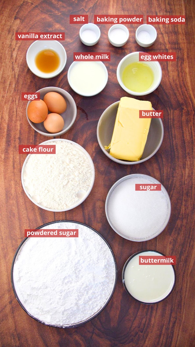 Top down view of ingredients used to make homemade vanilla cake with frosting including cake flour, eggs, sugar, baking powder, baking soda, salt, buttermilk, butter, milk, vanilla extract, and powdered sugar.