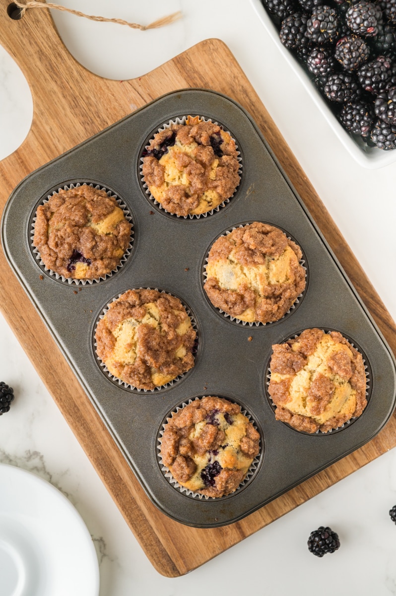 Blackberry Muffins, baked, and still in the pan, resting on a wooden cutting board, with blackberries in a white bowl on the side.