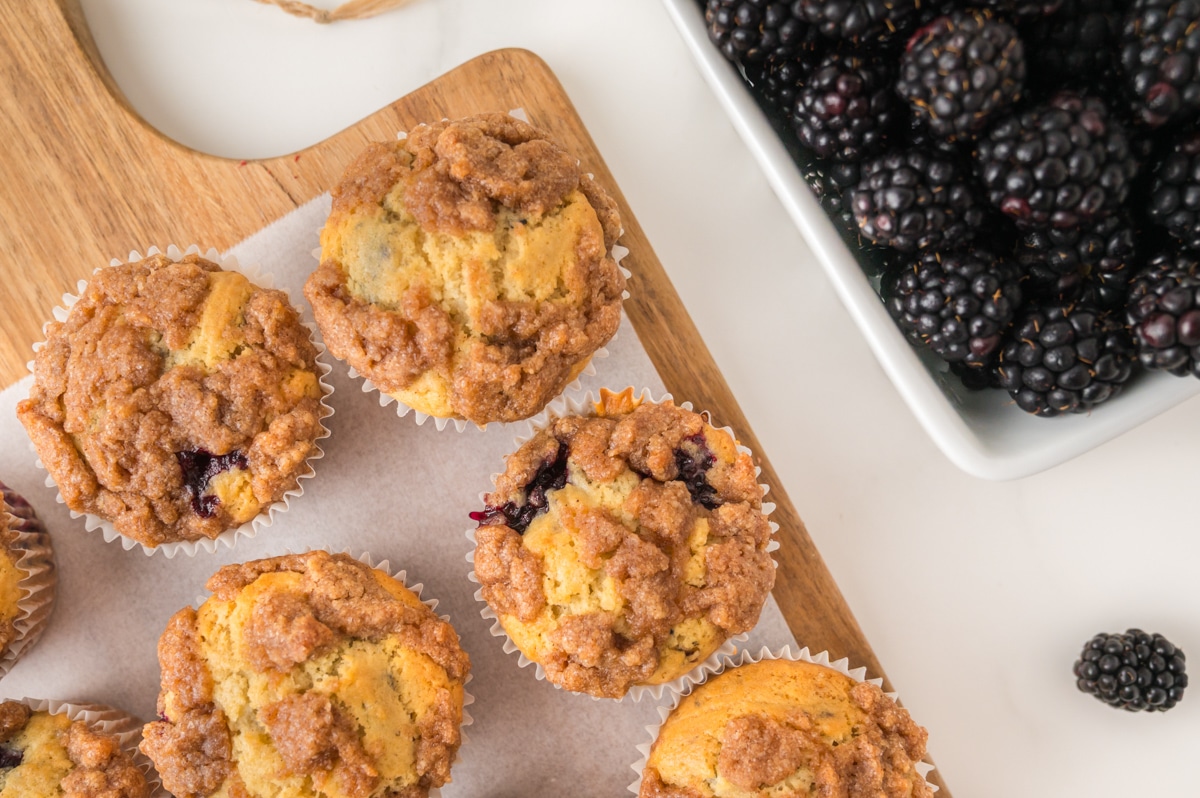 Blackberry Muffins cooling on a wooden cutting board with blackberries on the side in a white bowl.