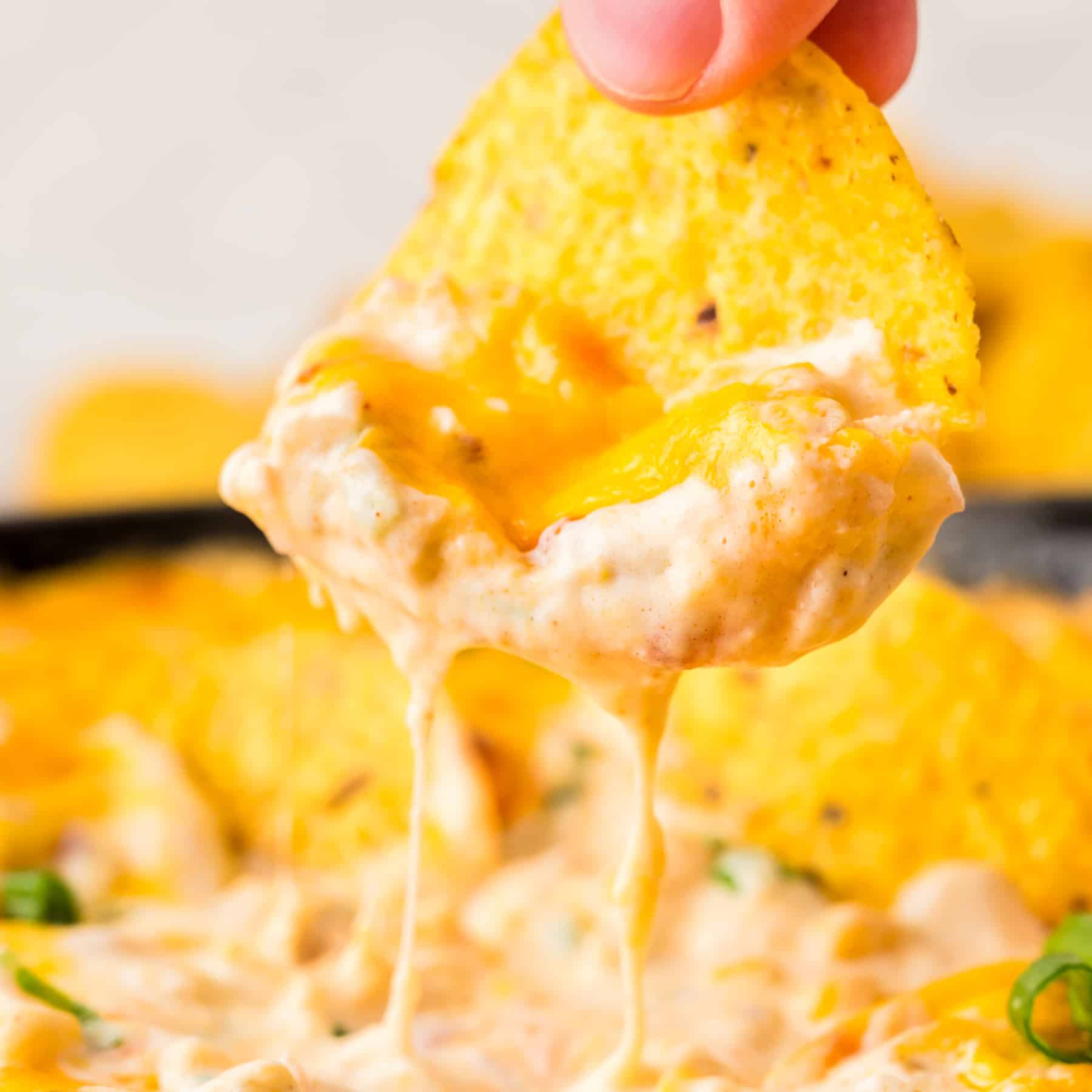 A hand holding a tortilla chip dripping with melted cheese and hot corn dip - Hostess At Heart