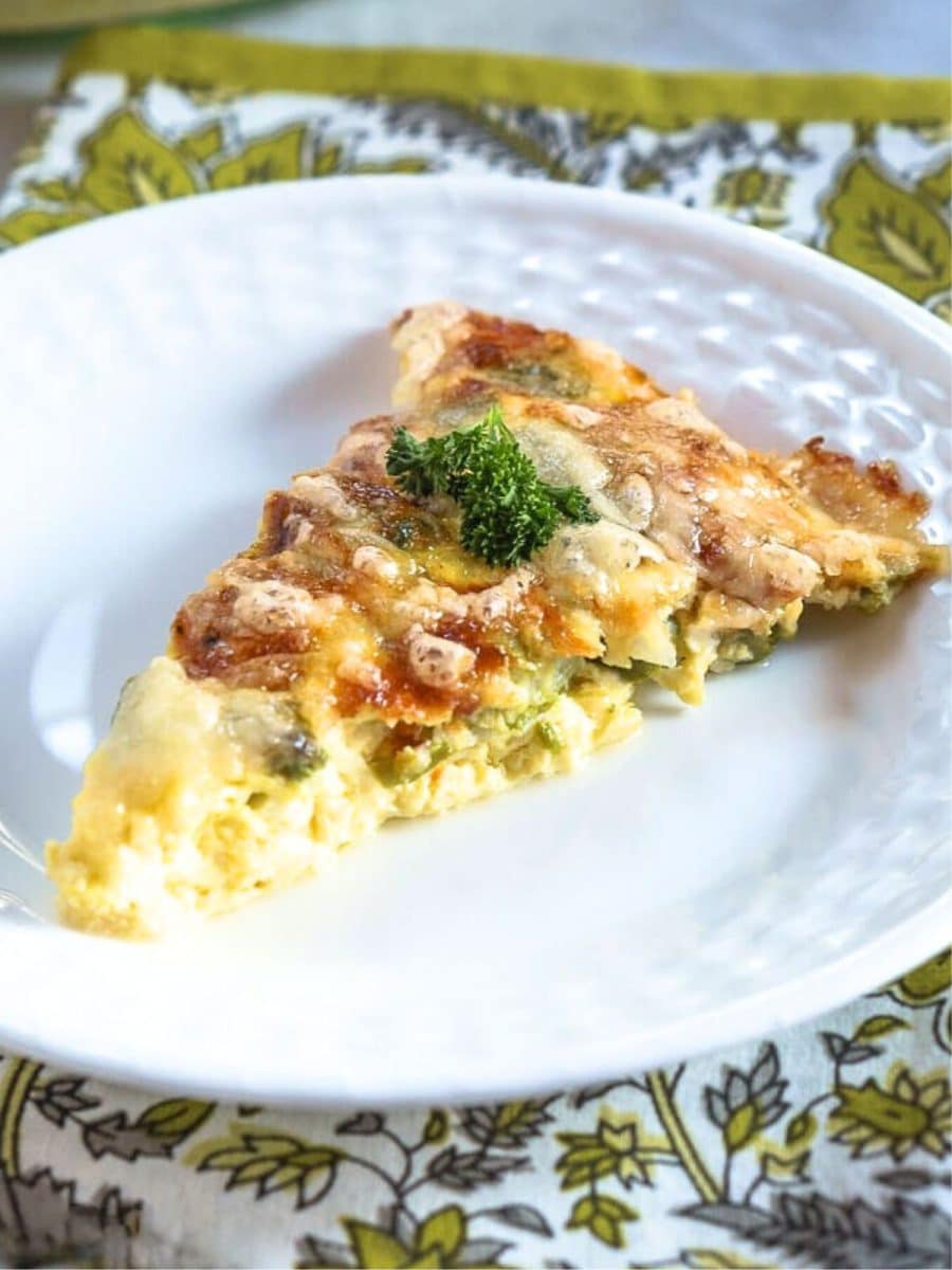 An angled view of a slice of asparagus quish topped with a sprig of parsley and melted cheese - Hostesss At Heart