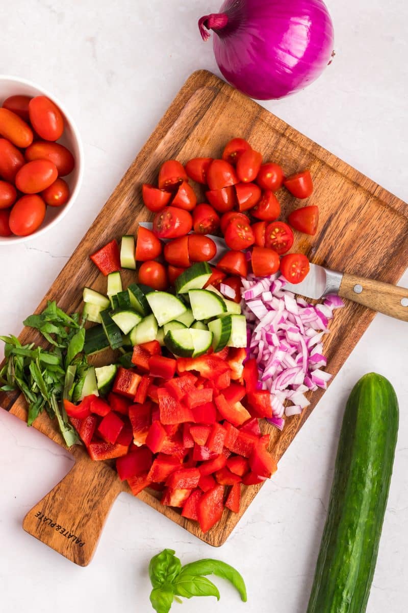 Top down view of a cutting board filled with ingredients necessary to make an Italian Tortellini Salad including red pepper, onion, cucumber, sweet basil, and tomatoes - Hostess At Heart
