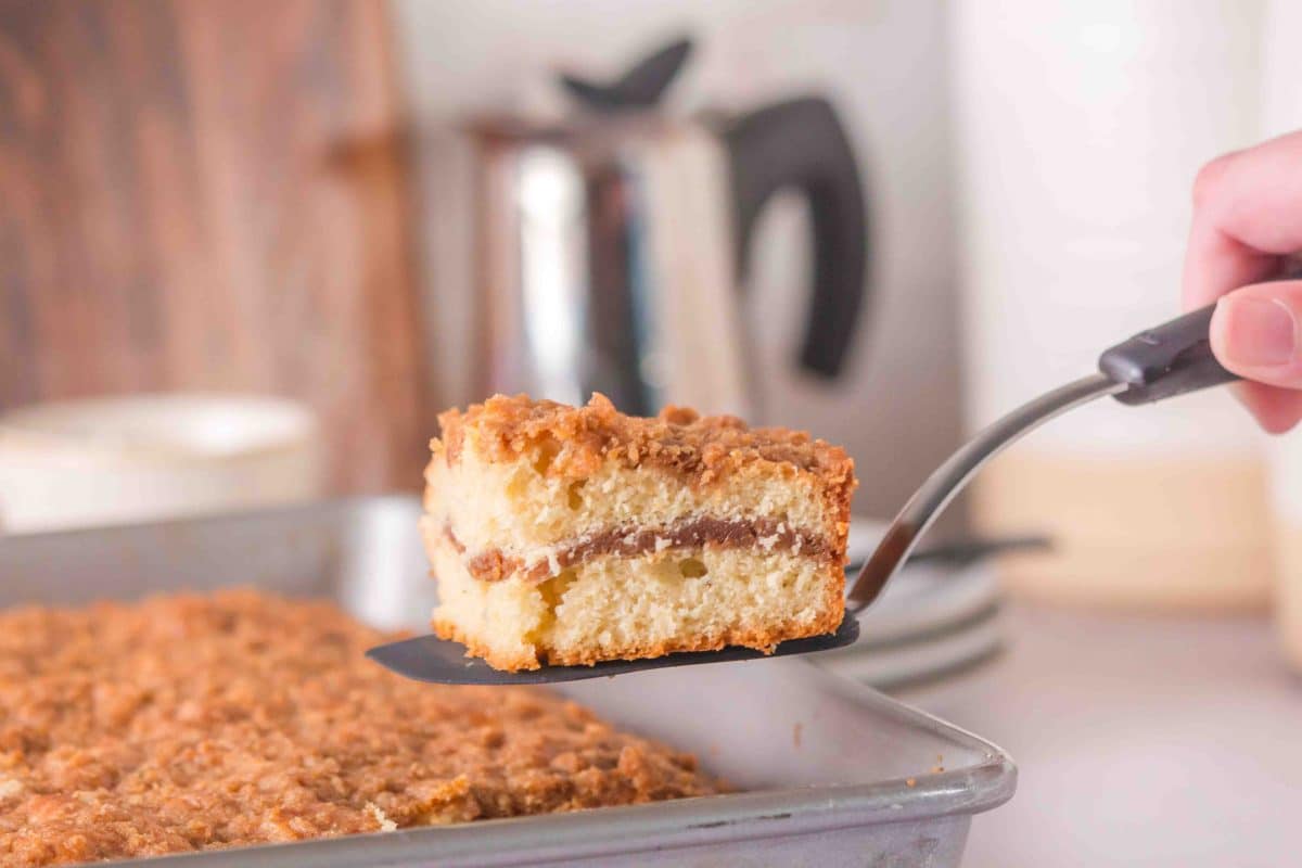 Close up of sourdough coffee cake on a spatula suspended over the pan with the whole cake in it, with a coffee carafe in the background.