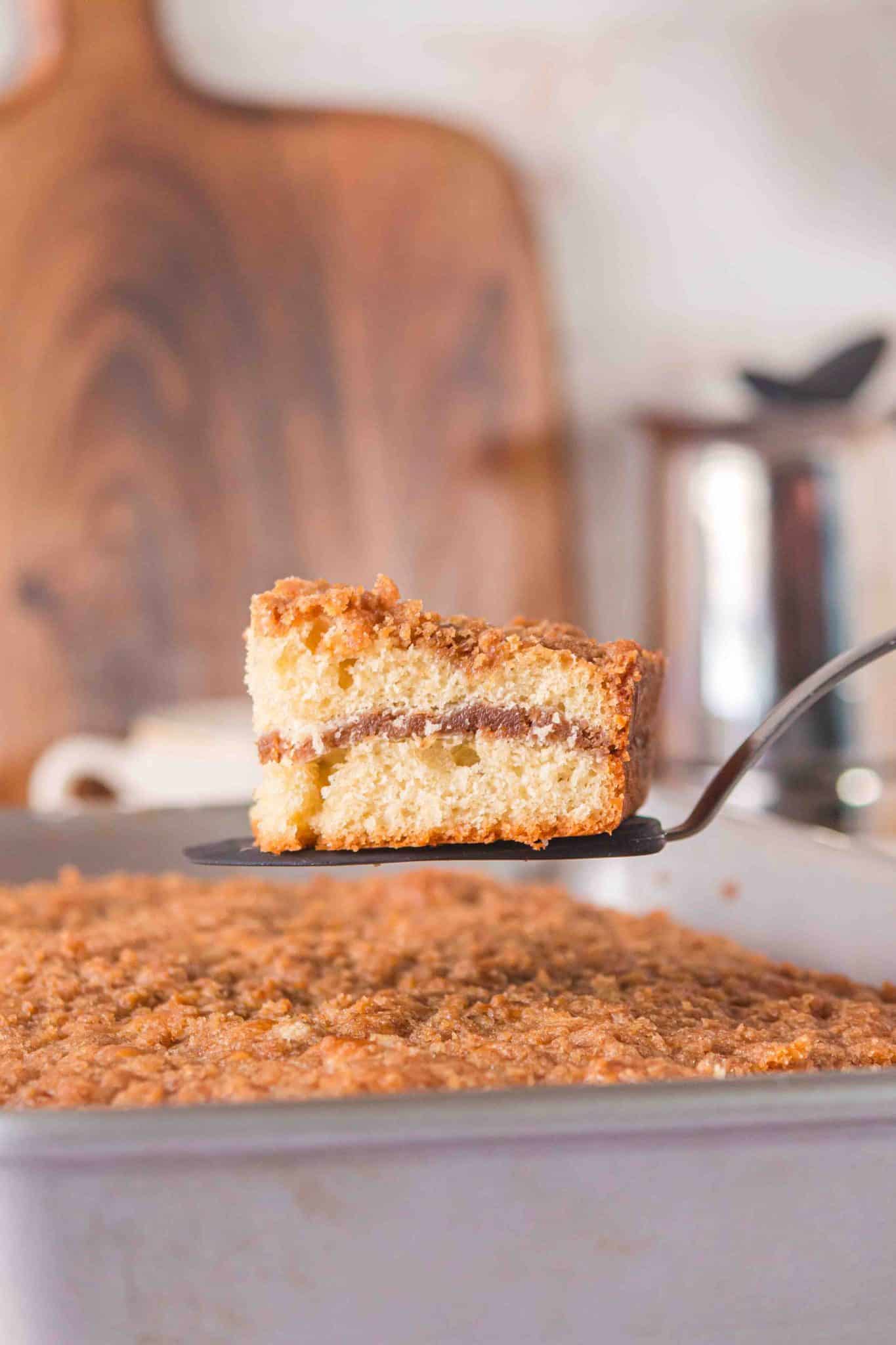 Close up side view of a slice of coffee cake suspended on a spatula over. the large cake with a coffee carafe and cutting board in teh background.