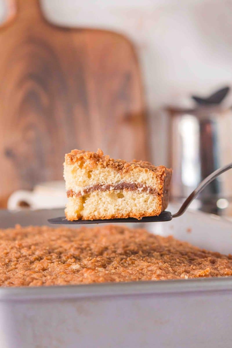 Close up side view of a slice of coffee cake suspended on a spatula over. the large cake with a coffee carafe and cutting board in teh background.